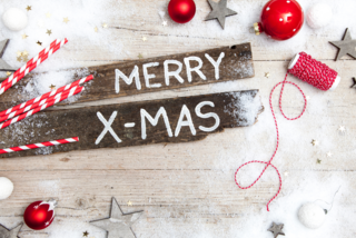 Weihnachtsgruß „Merry X-Mas“ in weißer Schrift auf zwei rustikalen Holztafeln, umgeben von Weihnachtskugeln, Sternen, Schneepulver und Geschenkband.